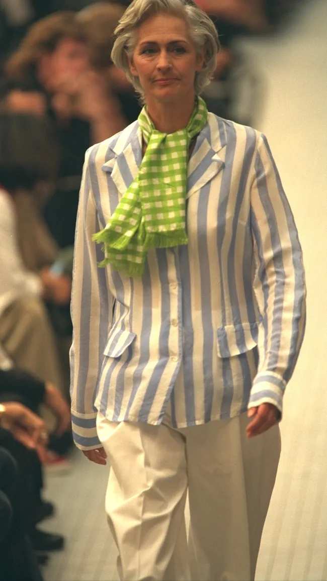 An older woman on a runway wears white pants, a blue-striped jacket, and a green checkered scarf.