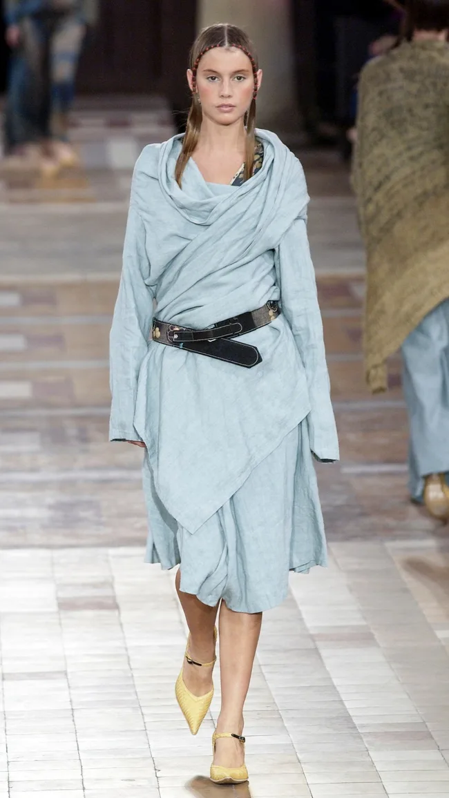 Model in light blue draped dress, black belt, yellow shoes, walking runway; minimalist fashion show setting.