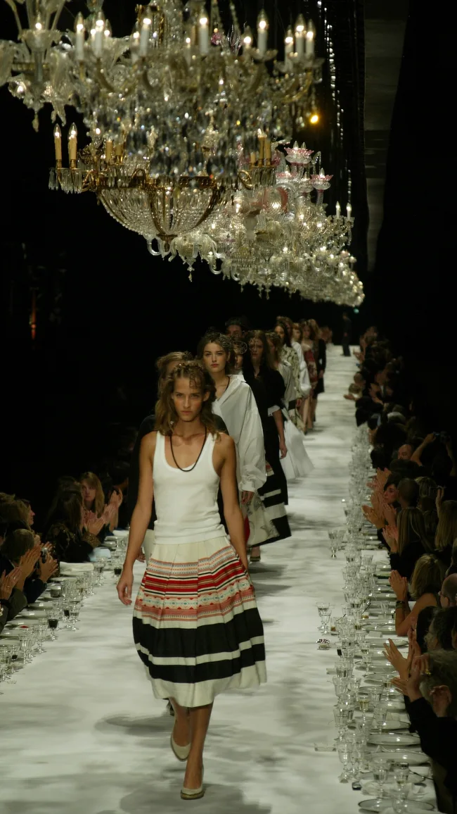 Models walking down a unique runway set as a long dining table with chandeliers above.