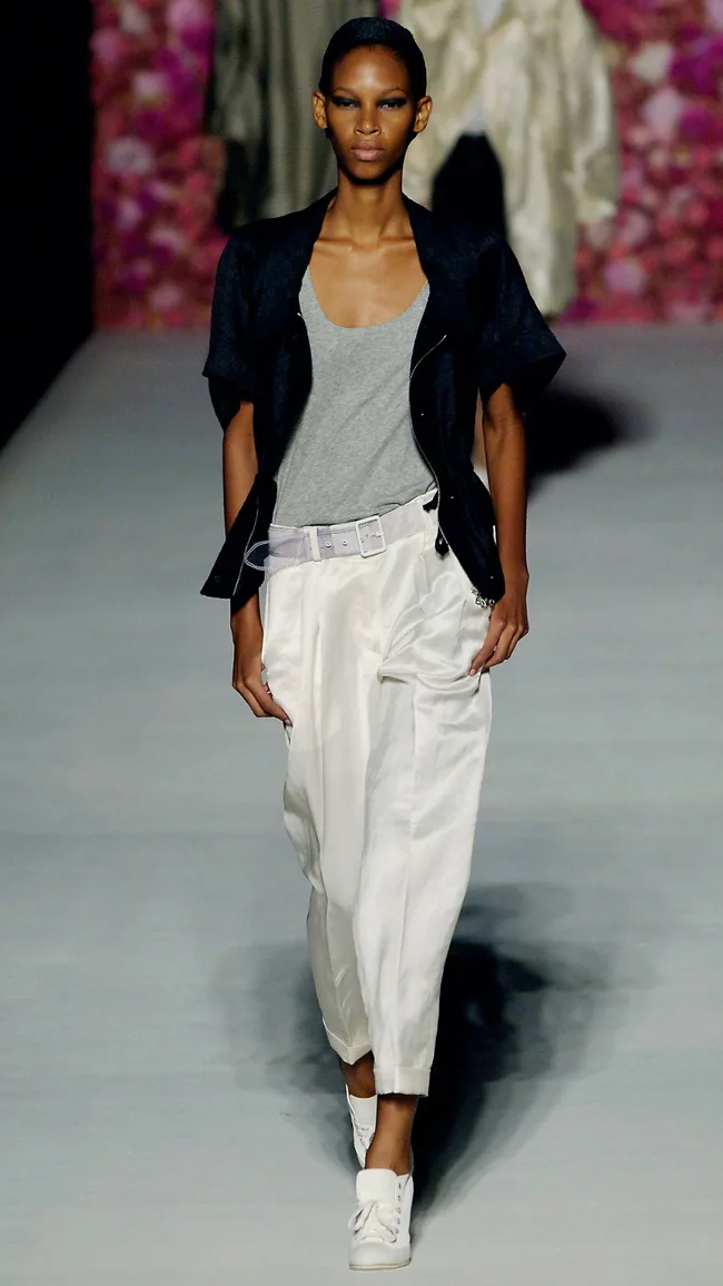 Model on runway wearing a grey tank, black jacket, white pants, and white sneakers against a floral backdrop.