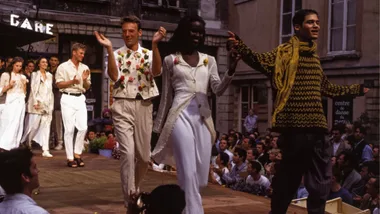 Models walking down a fashion show runway outdoors, with audience applauding in the background.