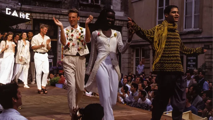Models walking down a fashion show runway outdoors, with audience applauding in the background.