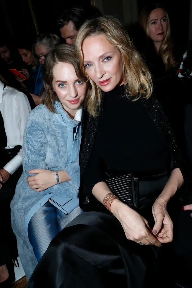 Two women sitting closely together, one wearing a light blue coat and the other in a black outfit, with people in the background.