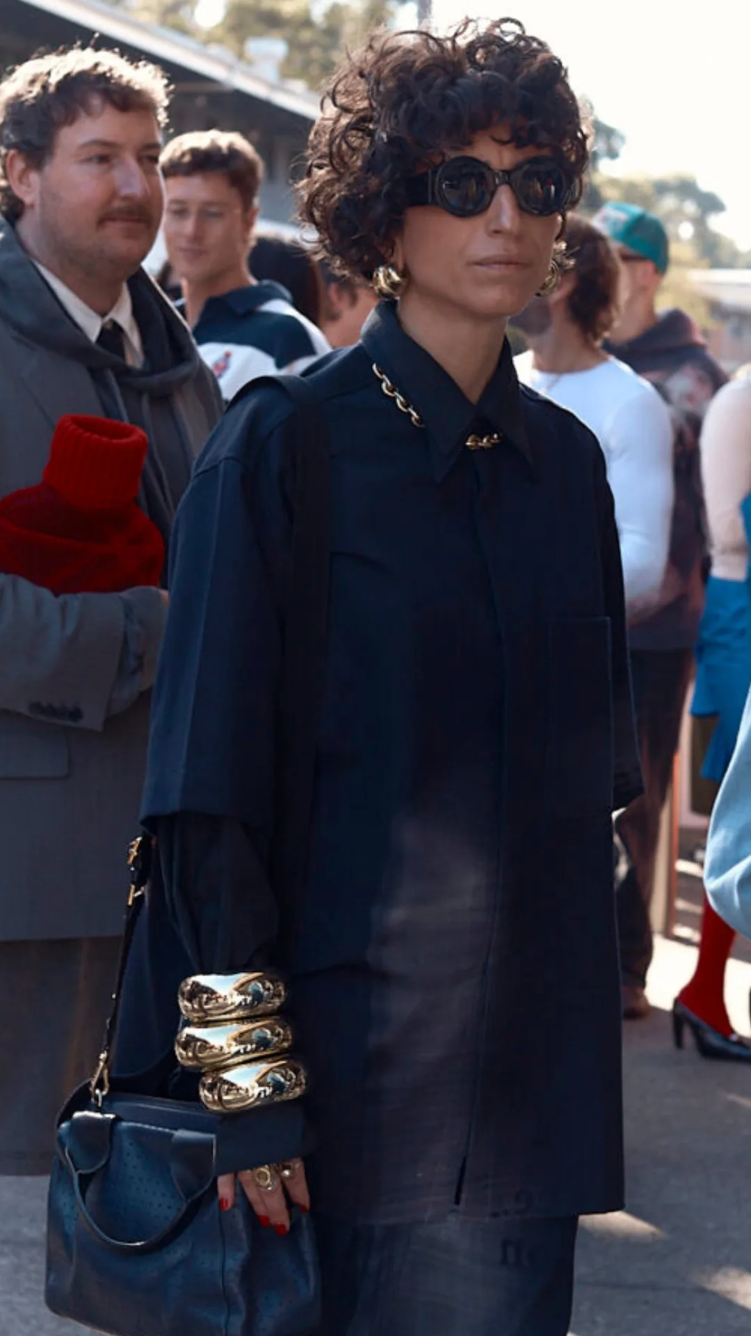 a stylish woman with short, curly black hair wears a black shirt and black pants. She wears black sunglasses and three big, gold bangles on top of her sleeve. She also wears gold earrings and a gold necklace, which peeks out from her shirt collar