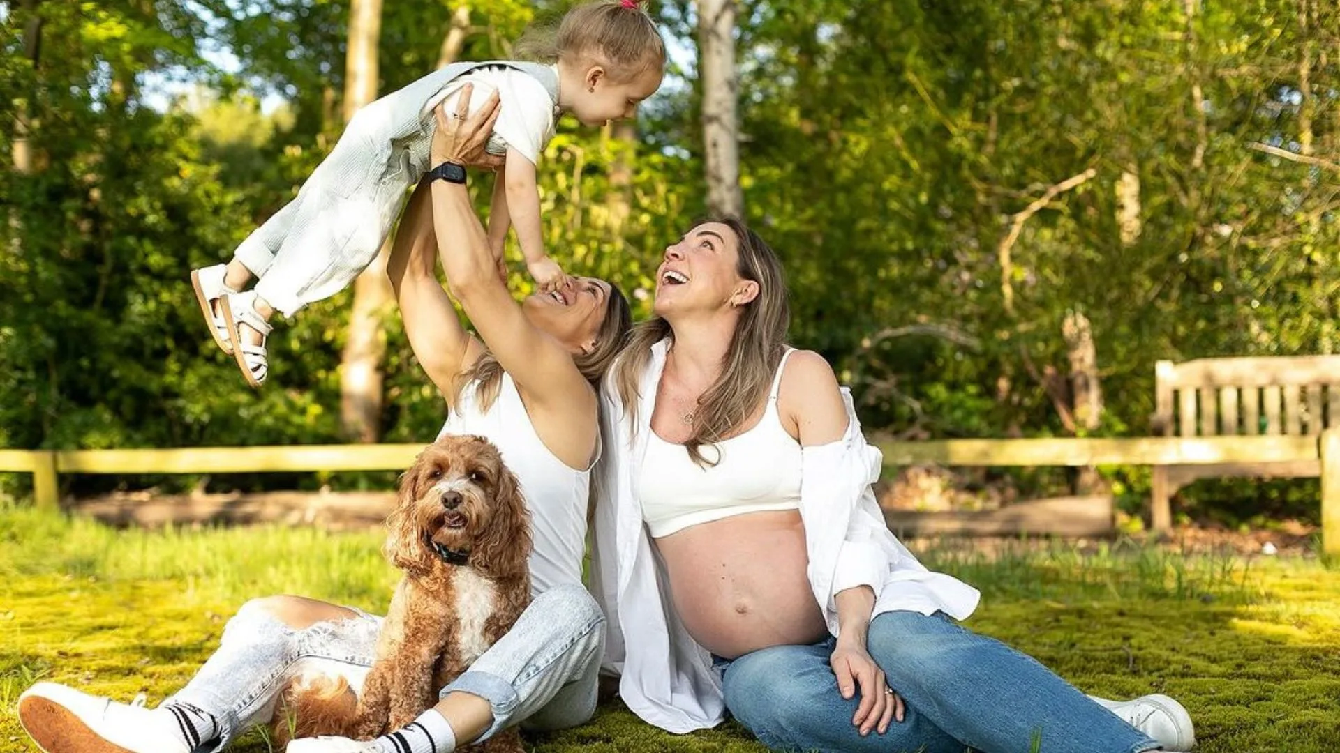 A happy family with two women, a child, and a dog sitting on grass in a sunny park.