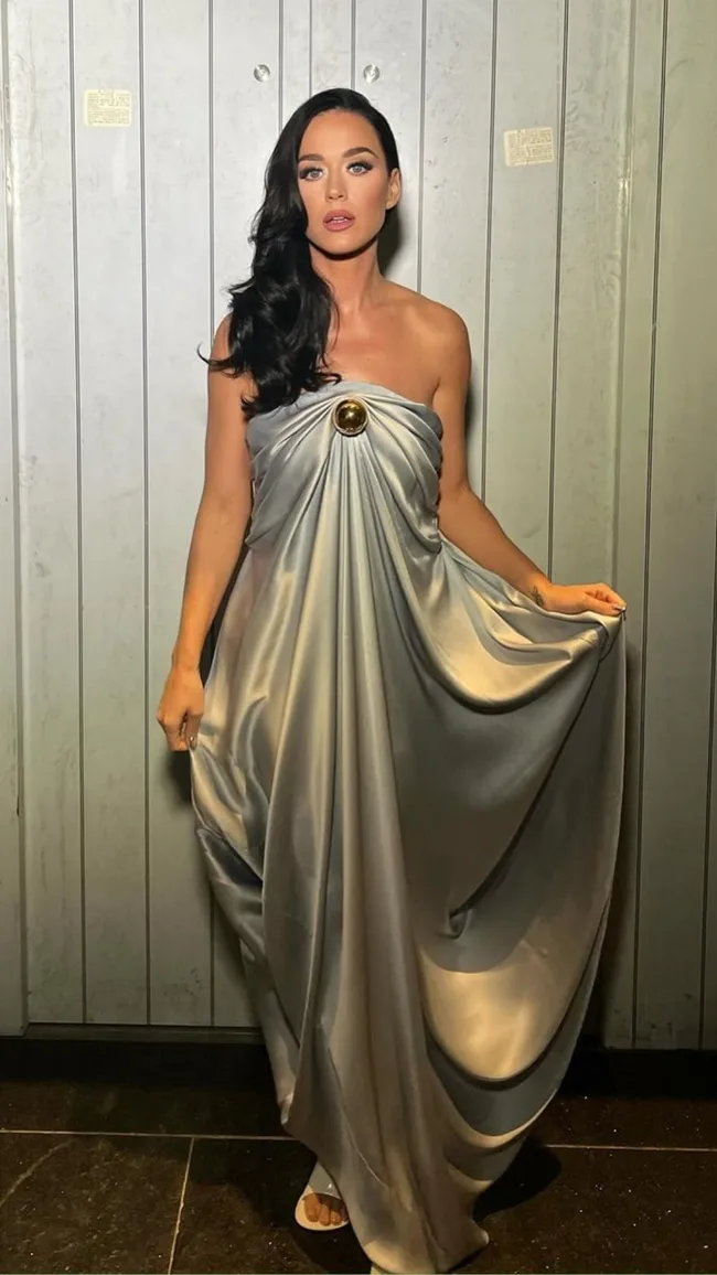 A woman in a silver gown with dark hair poses against a light panel background.