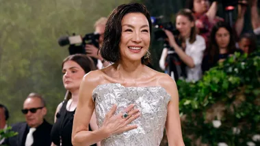 Person smiling in a textured silver dress at a public event, with onlookers and cameras in the background.