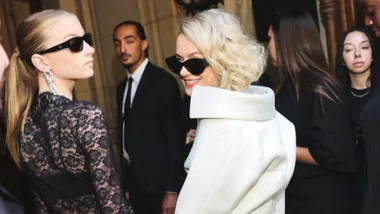 Two women in elegant outfits and sunglasses, attending a formal event, surrounded by other people.