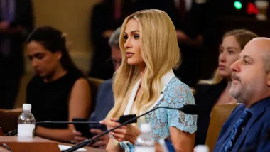 A person with long blonde hair in a light blue lace outfit sits attentively among others at a formal event.