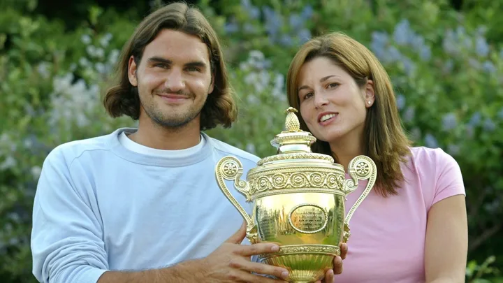 Two people smiling, holding a gold trophy with intricate designs, outdoors with foliage in the background.