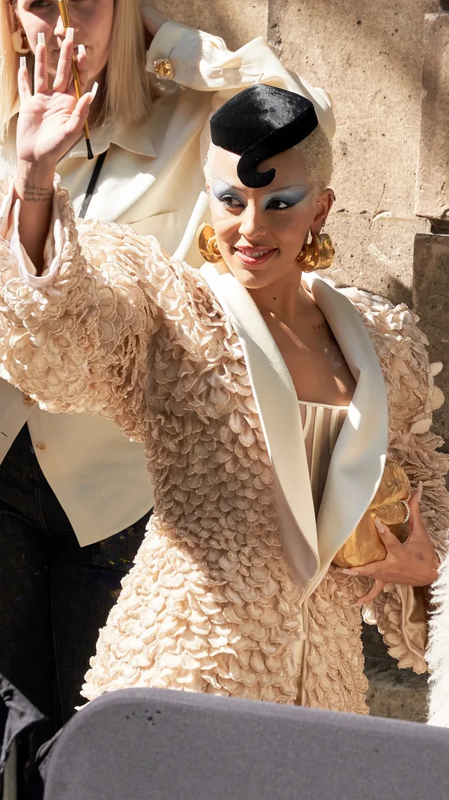 Woman in an elegant textured coat with bold makeup and a stylish hat, waving and smiling outdoors.