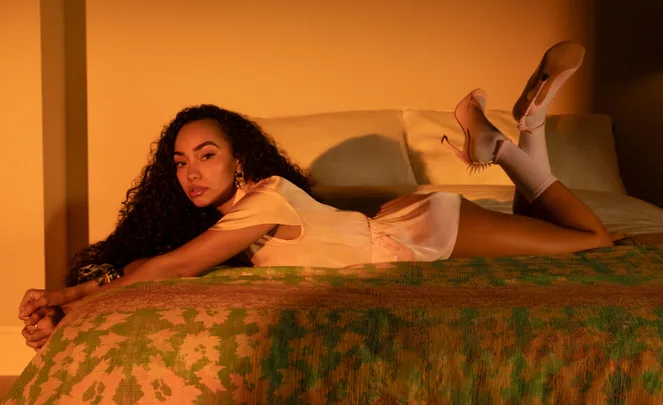 A person with curly hair lounges on a bed in pink attire and heels, set against a warm, softly lit bedroom.