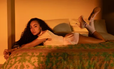 A person with curly hair lounges on a bed in pink attire and heels, set against a warm, softly lit bedroom.