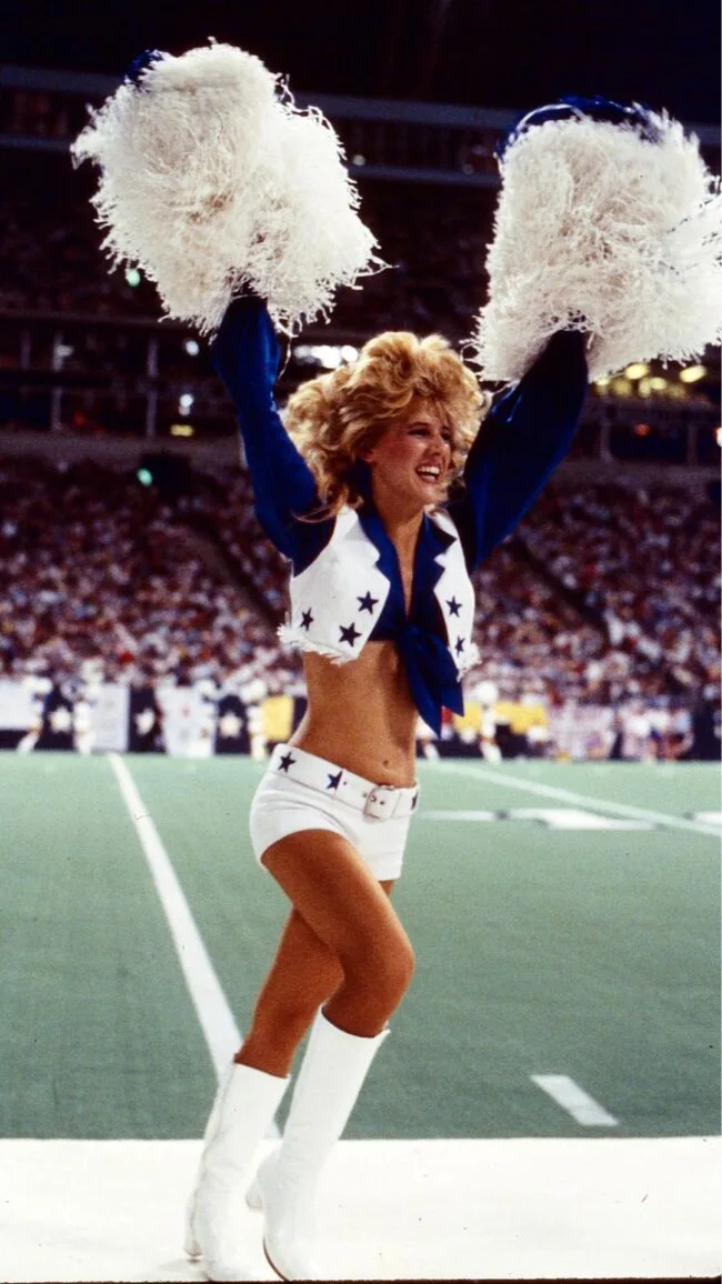 a blonde member of the dallas cowboys cheerleaders runs with pom poms above her head; her uniform consists of a blue shirt, white vest, small shite shorts and white gogo boots