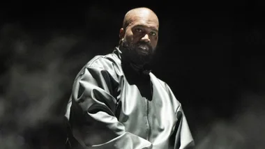 kanye west stares off-camera through a thin veil of smoke. He wears a vinyl bomber jacket, which is zipped up, and stands against a black background