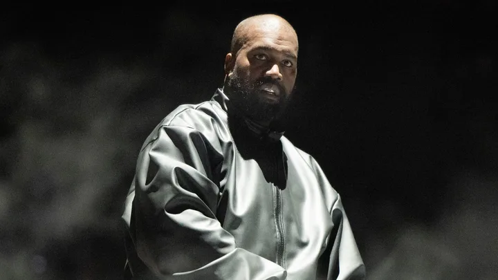 kanye west stares off-camera through a thin veil of smoke. He wears a vinyl bomber jacket, which is zipped up, and stands against a black background