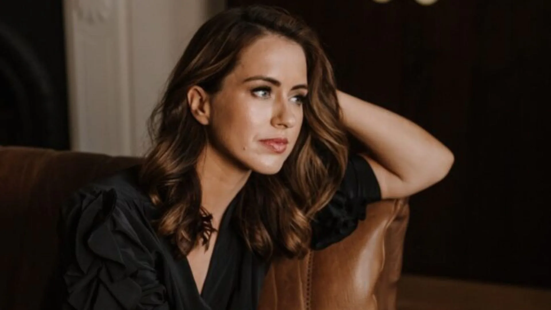 A woman with wavy brown hair is sitting on a couch, wearing a black outfit, looking contemplative.