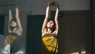 A woman with curly hair stretches post shower, wrapped in a yellow towel with a smile on her face.