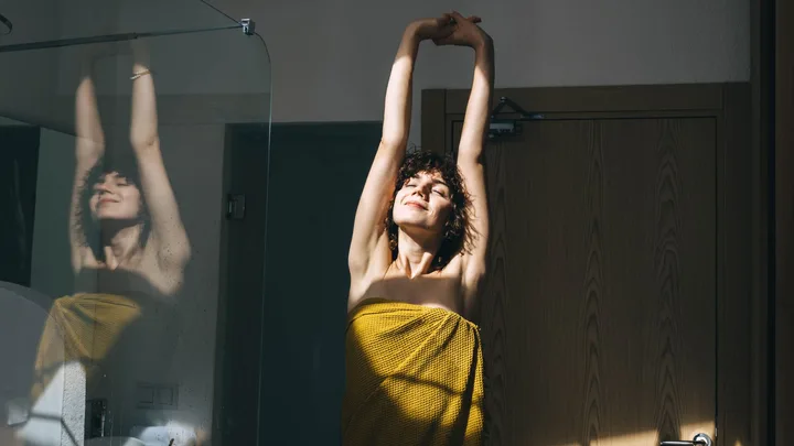 A woman with curly hair stretches post shower, wrapped in a yellow towel with a smile on her face.