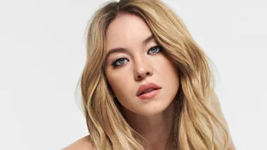 Portrait of a woman with long blonde hair, wearing natural makeup, gazing directly at the camera against a plain background.