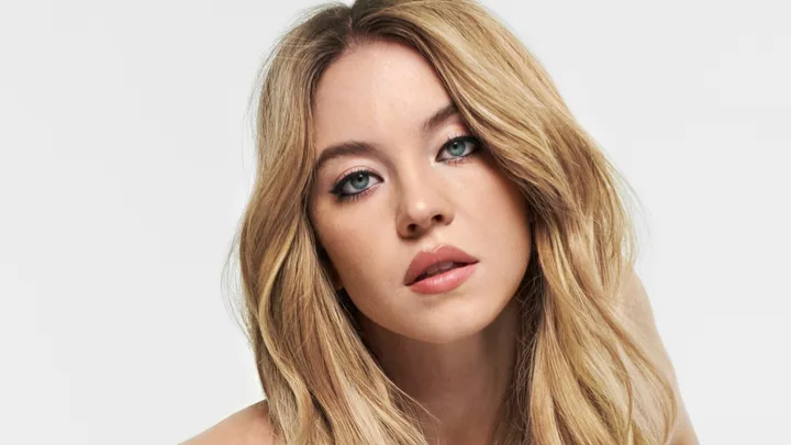 Portrait of a woman with long blonde hair, wearing natural makeup, gazing directly at the camera against a plain background.