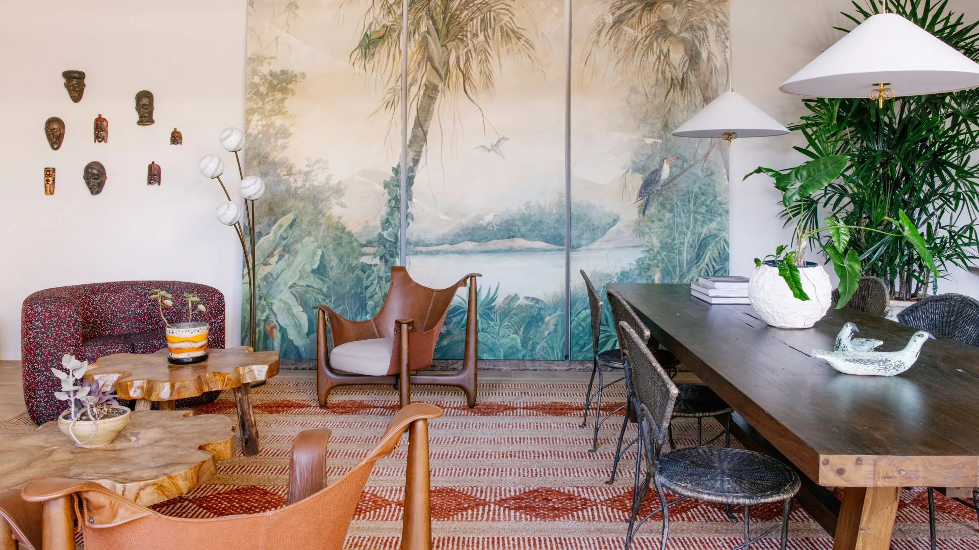 A living room in Basq House Byron Bay luxury accommodation with a purple armchair, leather chair and dark timber dining table with white cone-shaped pendant lights above it. On the back wall there is a wallpaper with a palm tree motif.