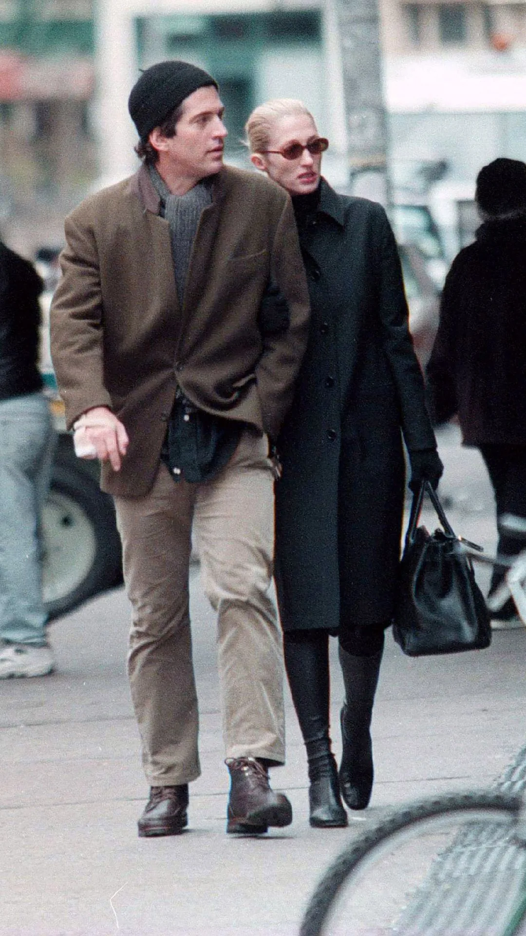 John F Kennedy Jr. and his wife Carolyn Bessette in New York. Carolyn is dressed in an all-black outfit and John is dressed in beige chinos with a brown blazer and black beanie. 