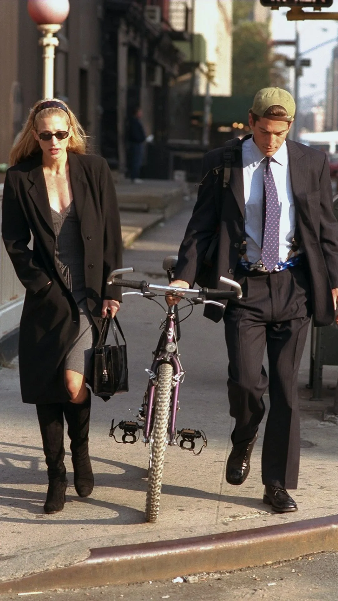 John F Kennedy Jr. and Carolyn Bessette Kennedy walk along Varick St New York. Carolyn is wearing a grey dress and black knee high boots and John is wearing a business suit with a green baseball cap, carrying his bike