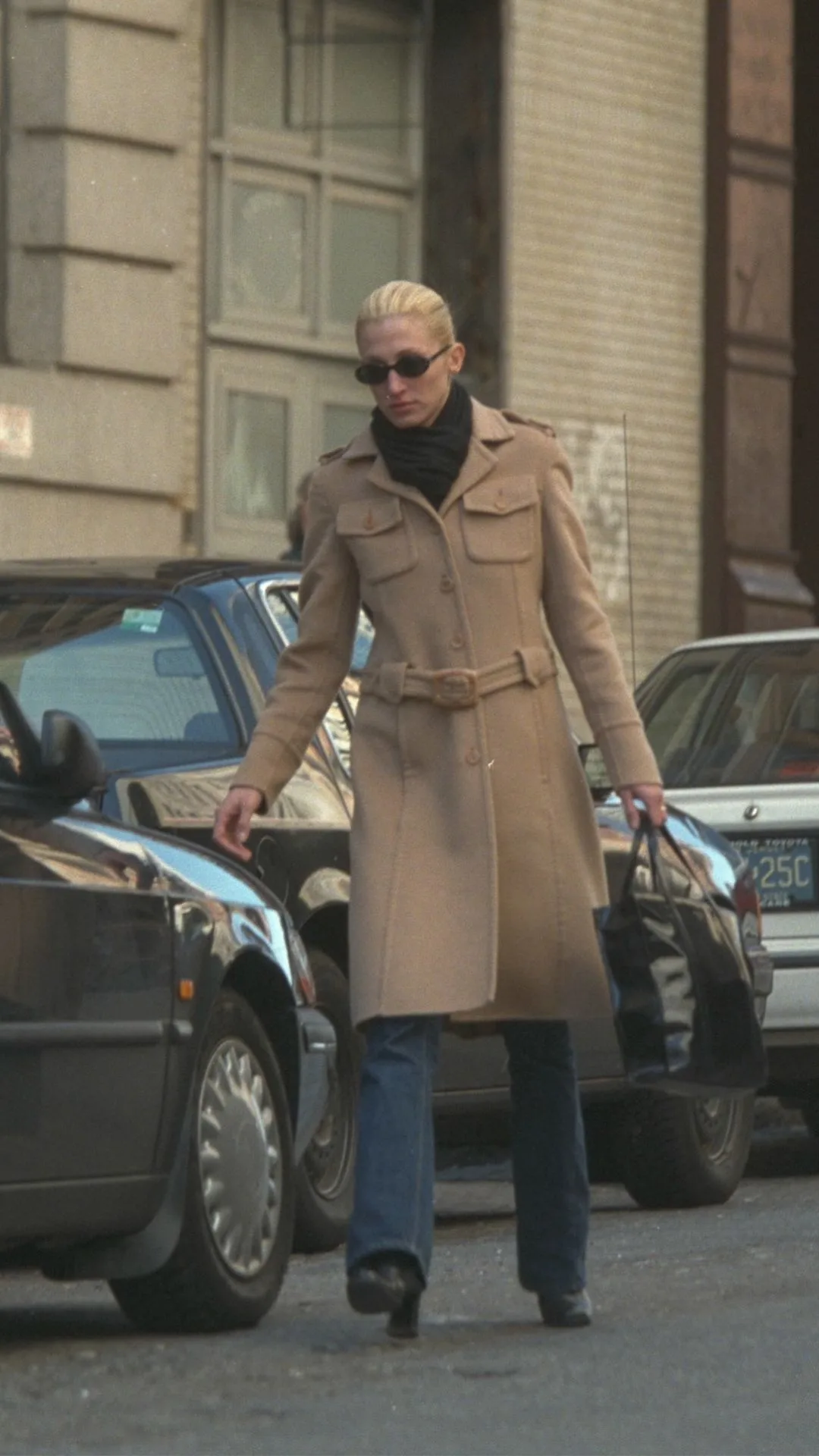 Carolyn Bessette Kennedy in a camel coat over blue jeans walking towards a car