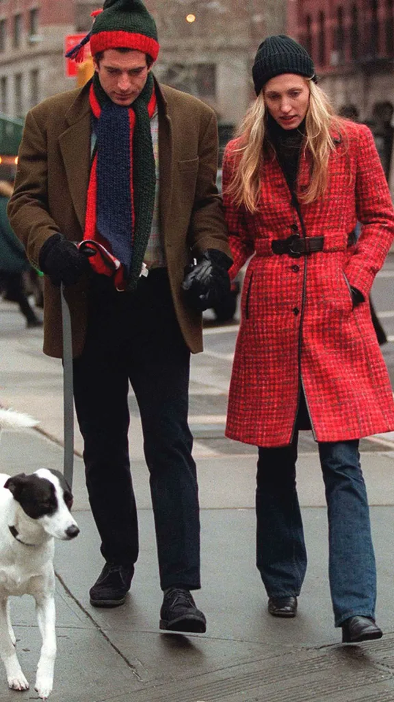 John F. Kennedy Jr. and his wife Carolyn walk with their dog. Carolyn is wearing a red checkered coat and jeans and John is wearing a green and red beanie and matching scarf.