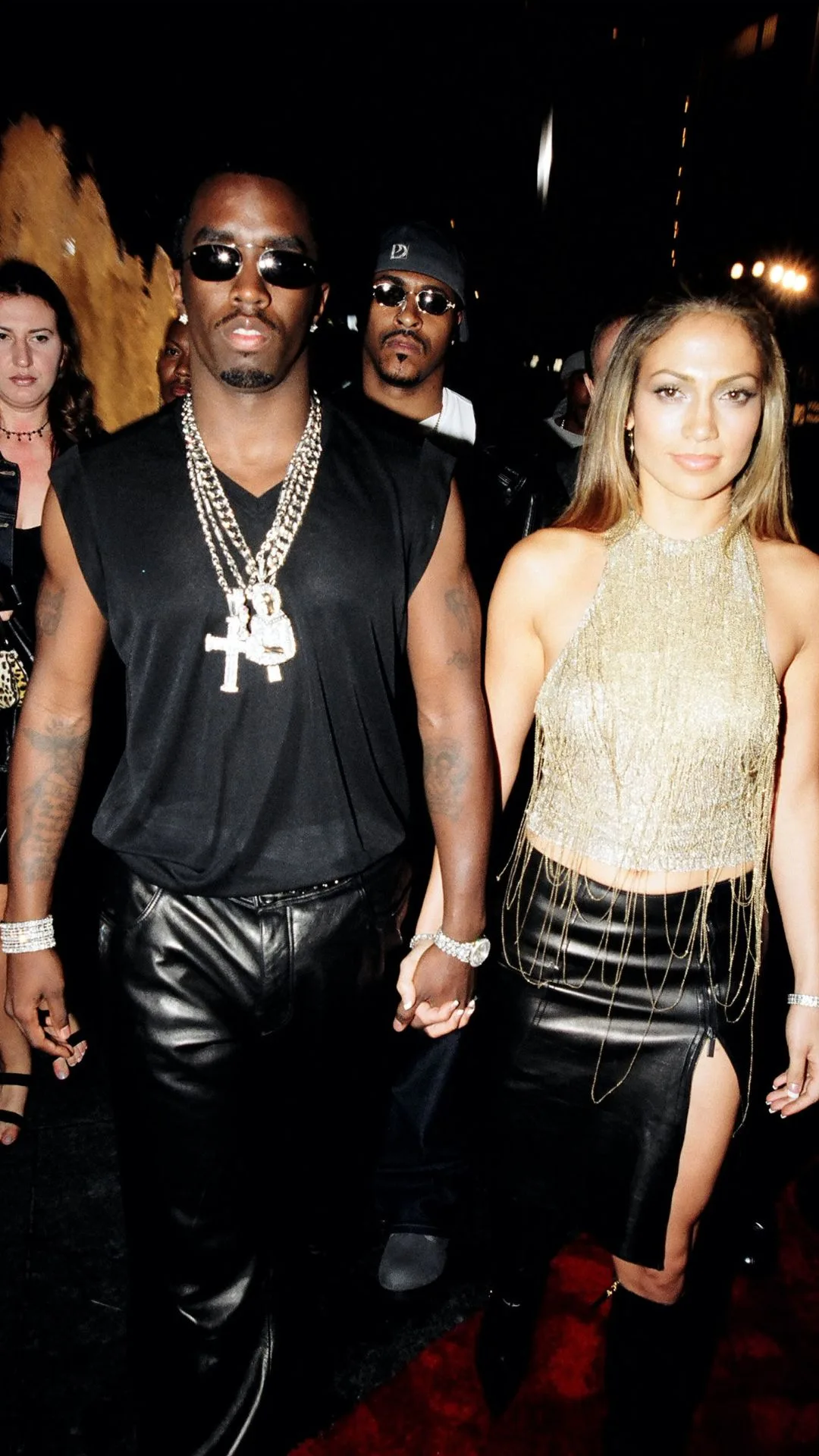 Sean Combs is wearing  series of silver diamond chain necklaces over a black t shirt vest and black leather pants and is holding hands with girlfriend Jennifer Lopez who is wearing a gold halterneck top and leather skirt. 