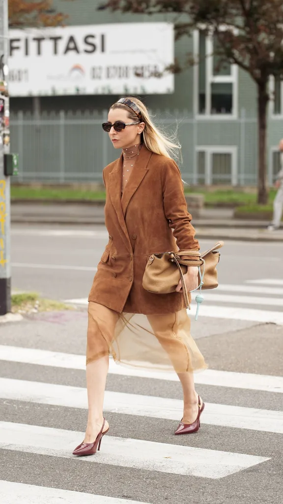 A woman crossing the road wearing a long tan suede blazer and sheer midi skirt at Milan Fashion Week