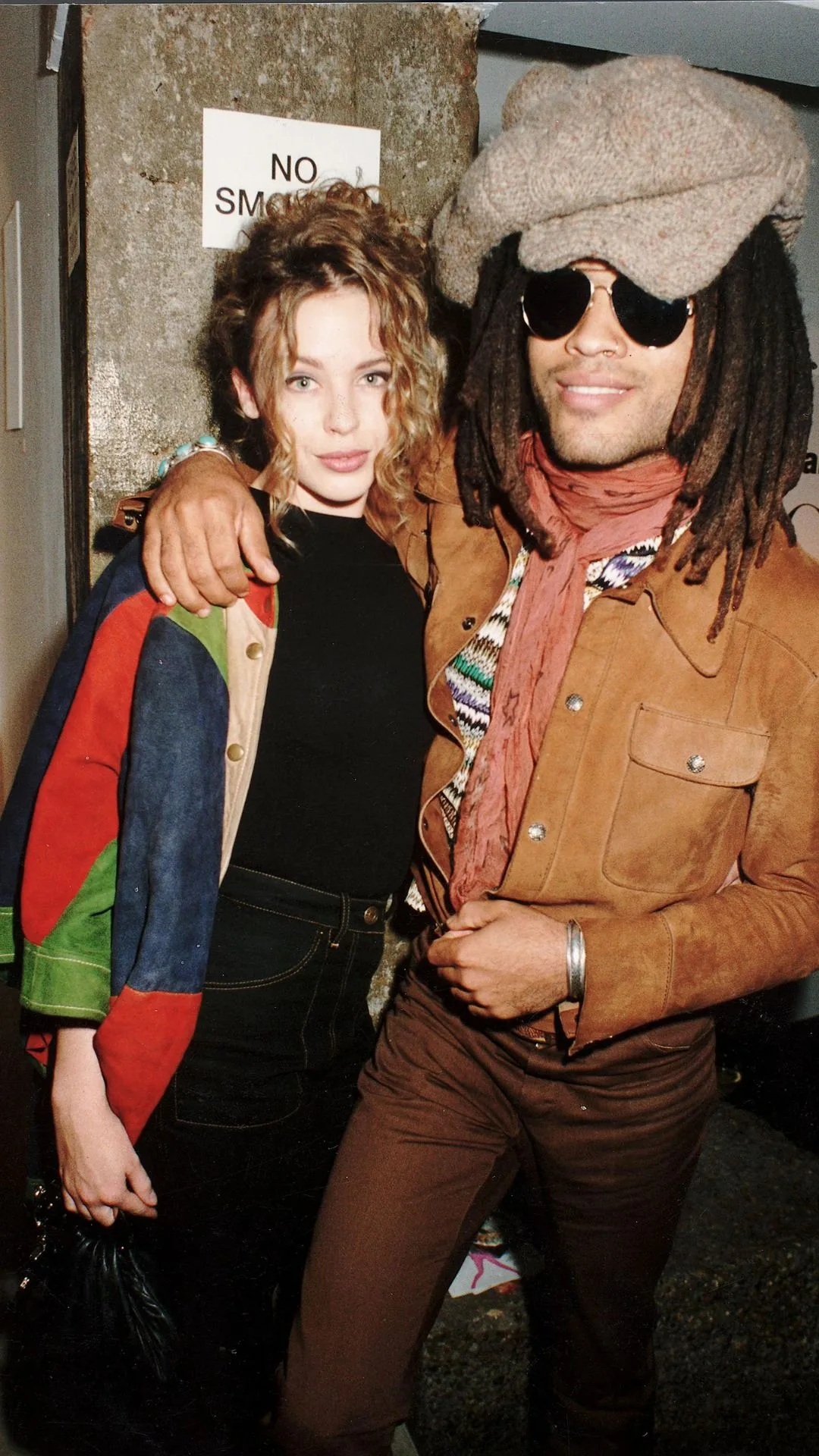 Singers Kylie Minogue and Lenny Kravitz at Vogue party in1995 in London. Kylie is wearing a black t shirt and dark wash jeans with a blue, green and red patterned denim jacket flung over her shoulder. Lenny is wearing a large knitted hat, sunglasses and a brown suede jacket and brown pants.