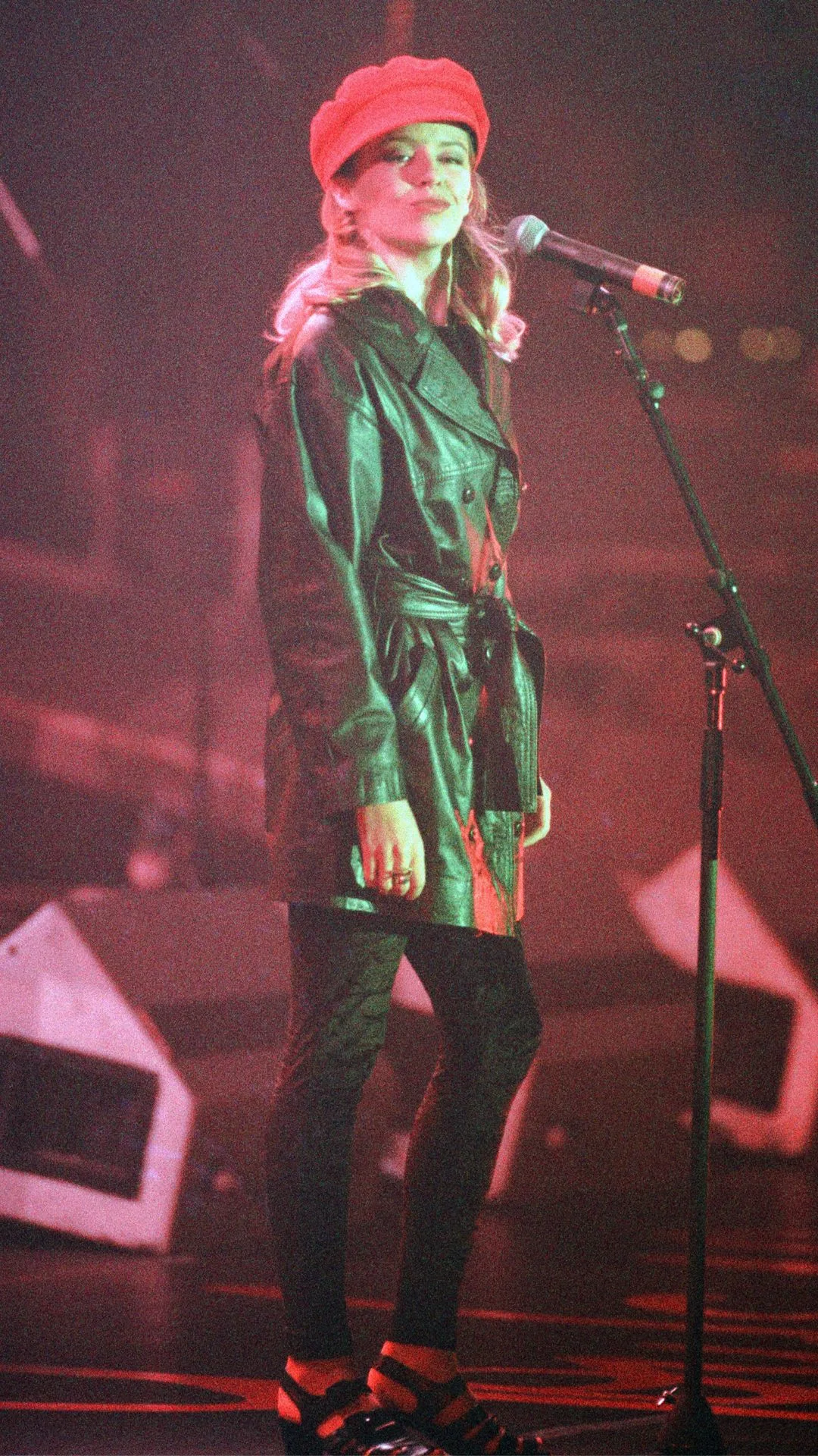 Kylie Minogue, performing in concert, Enjoy Yourself Tour, La Cigale, Paris. Kylie is on stage wearing a red beret, black stockings, and a cropped black leather trench coat. 