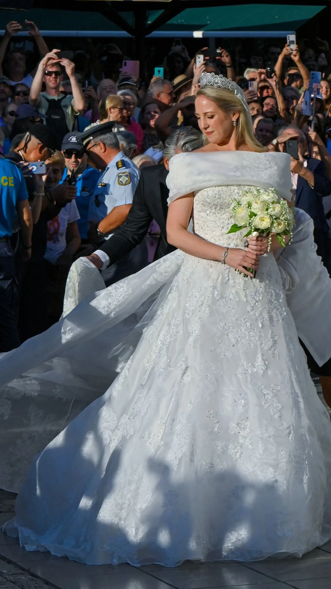 Princess Theodora of Greece arrives on her wedding day holding a bouquet of white roses and wearing a white off the shoulder embroidered dress. 