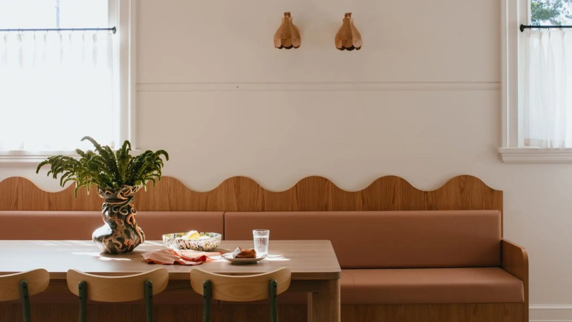 The dining area at Sona accommodation in Molong, NSW. A scalloped timber built-in bench seat with a pink leather upholstered seat sits behind the large oak dining table, set with a sculptural vase and plates. The back wall is painted in a warm white with two small scalloped wall sconces in between two windows covered in white cafe curtains.