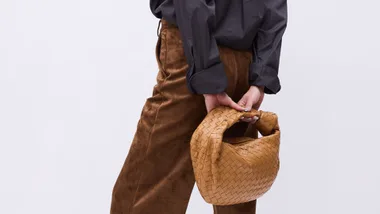 A close up of a woman holding a light brown handbag behind her back.