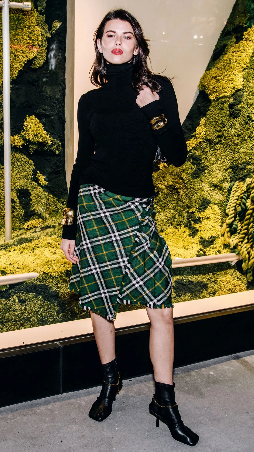 Harry Styles' ex-girlfriend Georgia Fowler attends the Burberry store reopening in New York in 2024. She is wearing a black turtleneck sweater and green and white tartan midi skirt with black square toe ankle boots. 