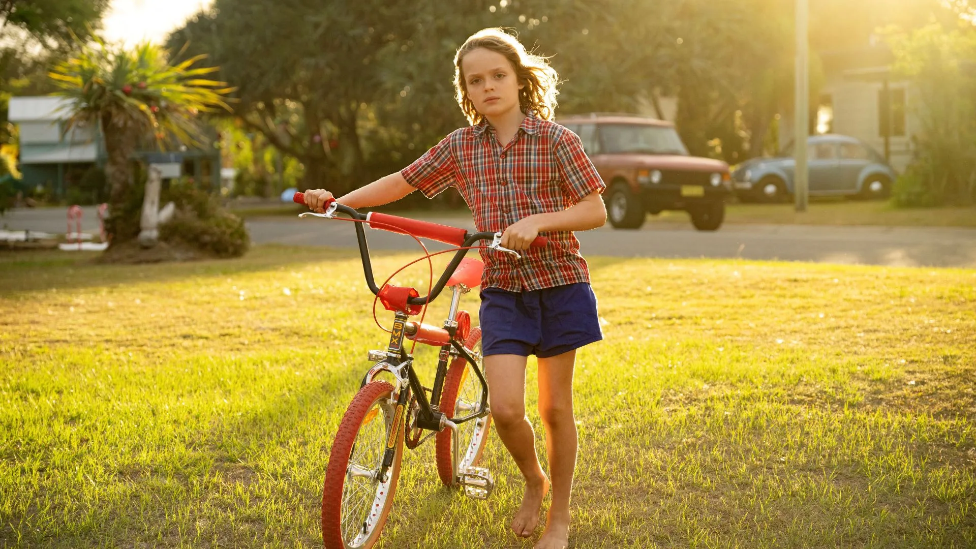 A film still from How To Make Gravy movie adaptation showing a boy in a garden walking next to his bike.