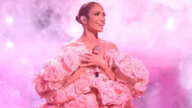 Musical guest Jennifer Lopez performs "This Is Me Now" on Saturday Night Live. The actress is seen on stage with pink smoke behind her as she holds a pink bedazzled microphone to her chest. She is wearing a pink puffy gown.