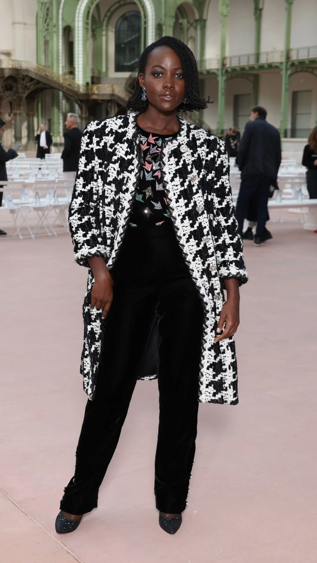 Lupita Nyong'o attends the Chanel womenswear show at Paris Fashion Week 2024 - dressed in black pants and a signature Chanel white and black herringbone coat