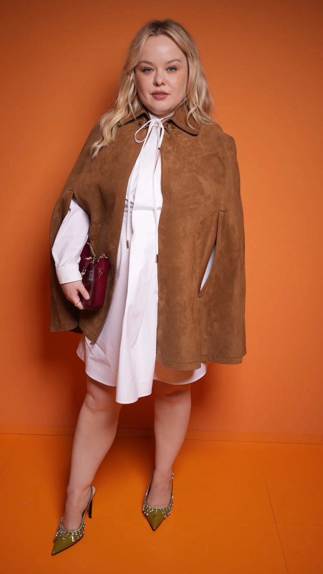 Nicola Coughlan at Gucci RTW Spring 2025 during Milan Fashion Week. The actress is up against an orange backdrop and wears a white shirt-style mini dress, with a brown suede cape over it. 