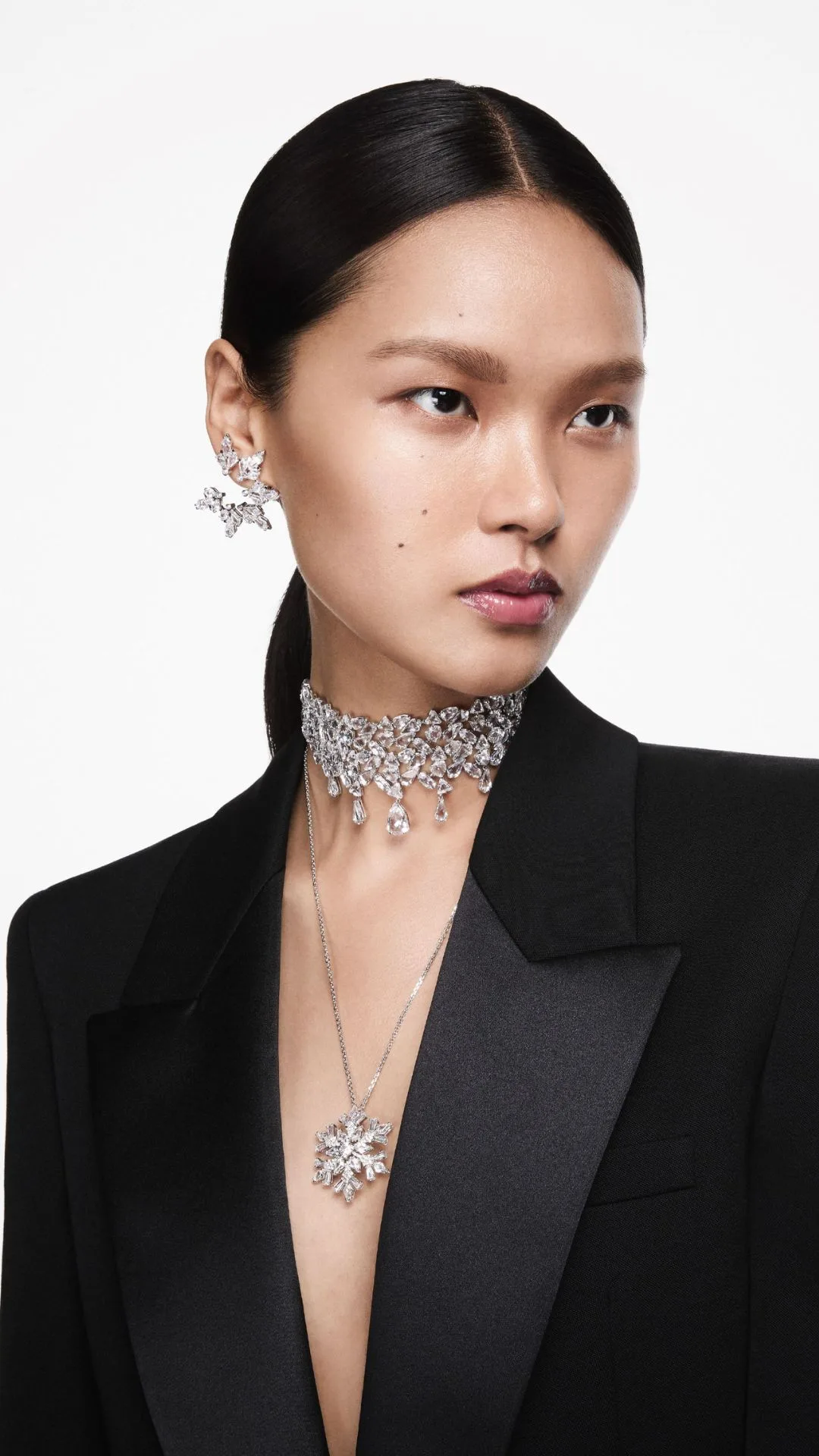 A model wearing a black tuxedo jacket models Swarovski's fall winter 2024 range. She wears a dazzling crystal choker and long pendant necklace with a crystal snowflake motif and matching crystal snowflake-like earrings.