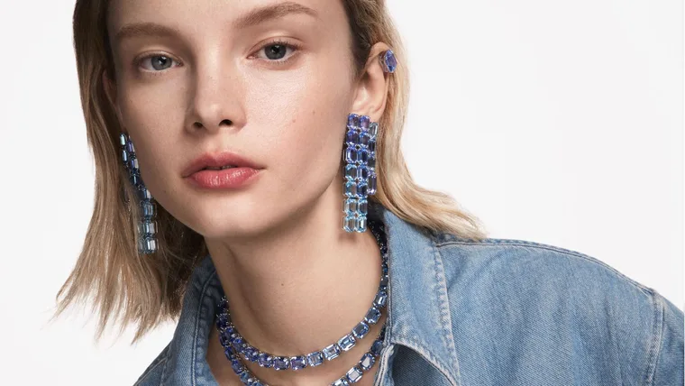 A close up of a blonde Caucasian model in a light denim shirt wearing statement Swarovski crystal pendant earrings in ombre blue colours, with matching ombre blue necklaces.