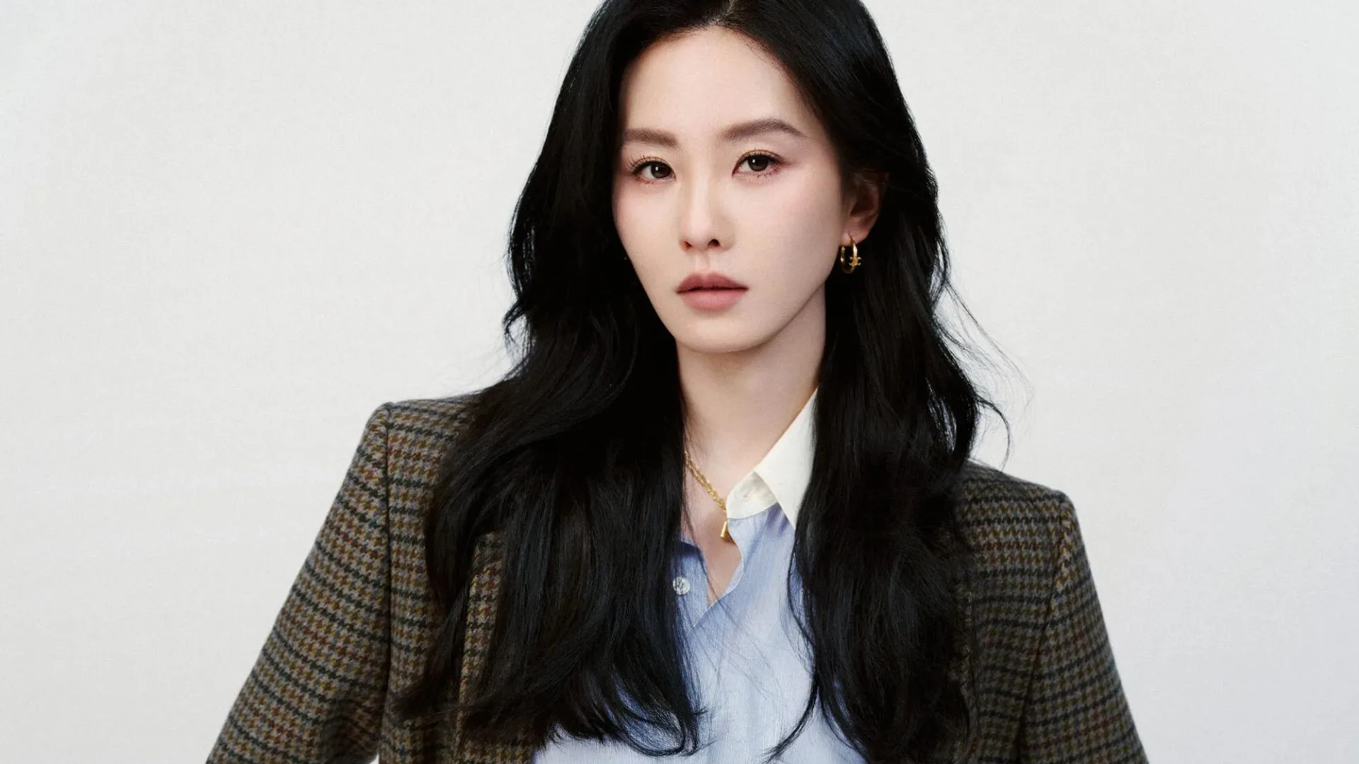 Portrait of a woman with long black hair, wearing a brown plaid blazer and light blue shirt, against a plain background.