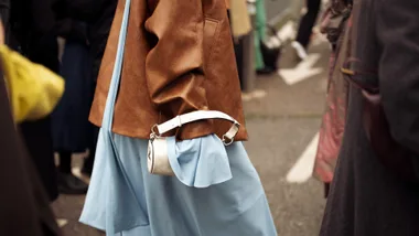 A guest wears a blue dress, brown leather jacket, and white bag outside the Gucci fashion show during the Milan Fashion Week