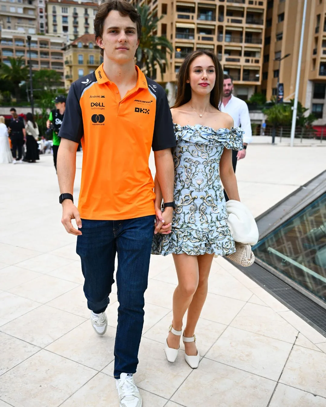 A man wearing an orange polo shirt and jeans walking hand in hand with a young woman wearing a blue and white floral dress. 