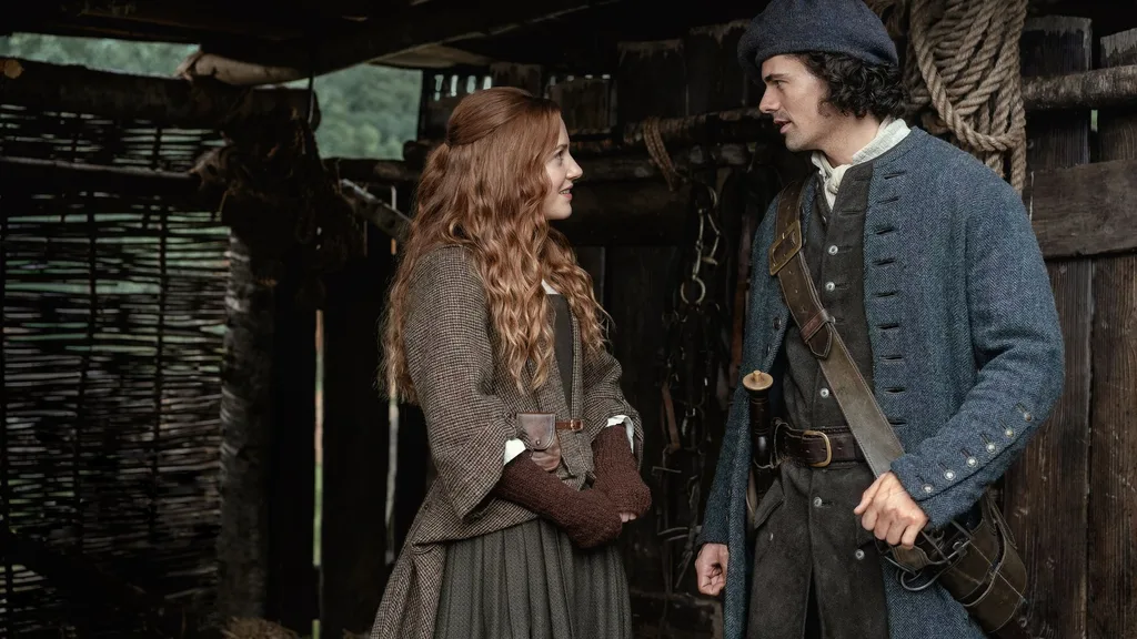A young woman with red hair talks to a young man weaing traditional Scottish dress outside a barn in 18th century Scotland.