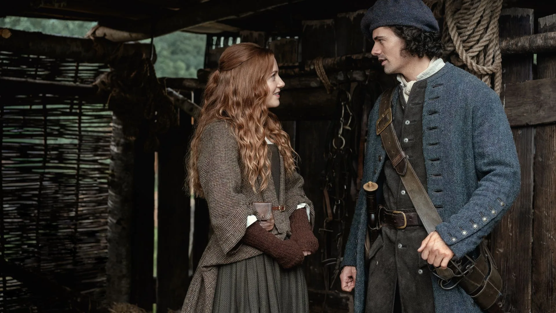 A young woman with red hair talks to a young man weaing traditional Scottish dress outside a barn in 18th century Scotland. 