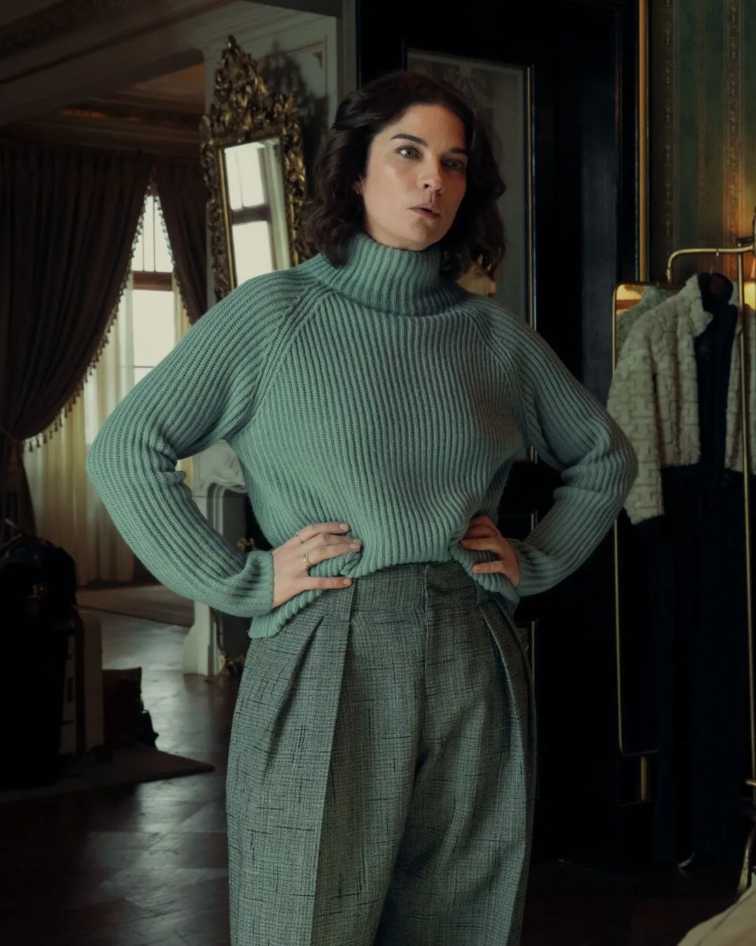 A brunette woman with short hair stands with her hands on her hips. She is wearing a duck egg blue turtleneck jumper and  grey tailored pants. 